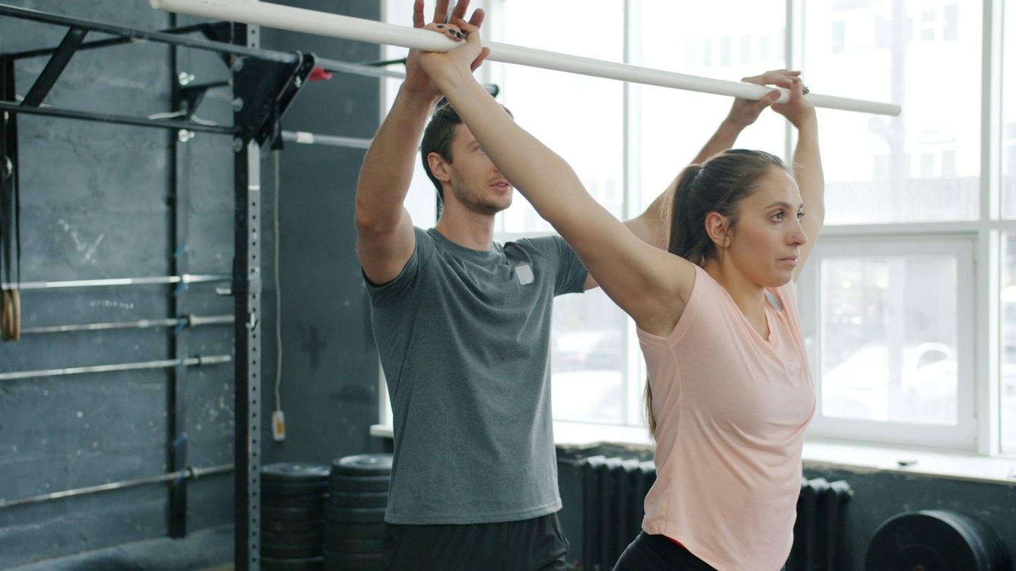 Trainer corrects woman's form with a bar in a gym.